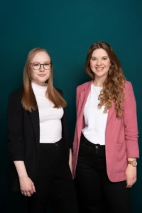 Two smiling women in blazers pose against a teal backdrop in a professional, studio-lit team portrait.