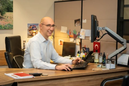 Smiling man in a white shirt and glasses seated at a wooden desk typing in a warm office, an authentic business headshot.