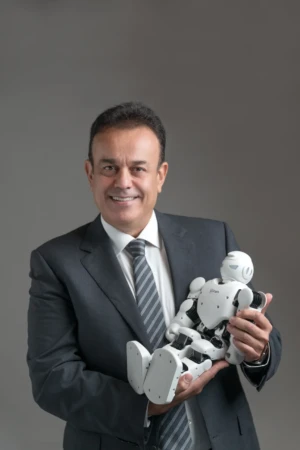 Smiling man in a dark suit holds a white robot against a grey backdrop with soft studio light, a modern executive portrait.