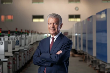Confident executive portrait of a gray-haired man in a navy suit with folded arms in a blurred industrial setting.