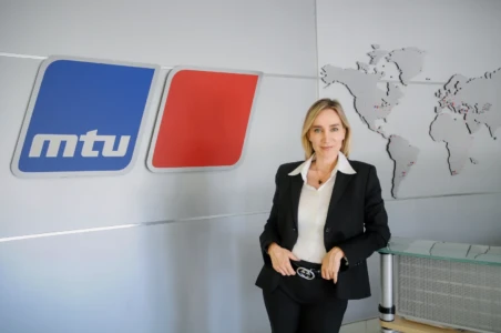 Confident woman in a black suit poses by an MTU logo and world-map wall in a professional corporate headshot.