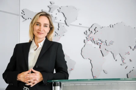 Confident corporate headshot of a smiling blonde woman in a black suit leaning on a glass desk against a world map.