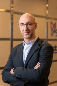 Confident corporate headshot of a bald man wearing glasses and a dark blazer, arms crossed, warm studio light.