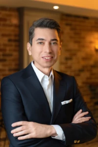 Smiling man in a dark suit with folded arms against a warm brick wall, confident professional headshot lit with studio light.