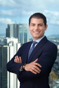 Confident corporate headshot of a smiling man in a navy suit with arms crossed against a blurred city skyline in soft light.