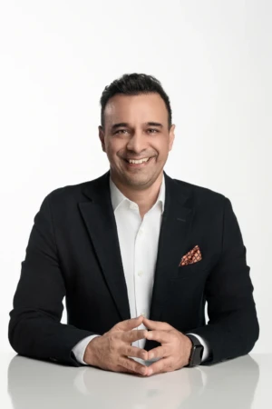 Smiling man in a dark blazer and white shirt, posed at a desk for a confident business headshot with clean studio light.