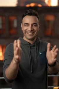 Smiling man in a dark hoodie reaching toward camera with blurred hands against warm bokeh, authentic personal branding photo.