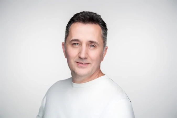 Smiling middle-aged man in a white shirt against a neutral backdrop, soft studio light, professional headshot.