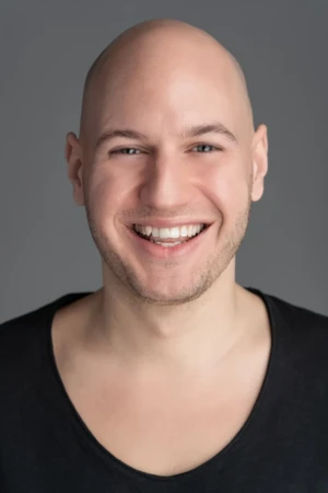Warm, smiling bald man in a black shirt against a neutral gray backdrop, studio-lit authentic professional headshot.