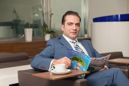 Confident man in a blue suit seated in an office lounge reading a magazine with a coffee, professional business headshot.