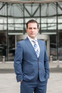 Man in a blue suit with patterned tie stands before a glass office building, confident expression, business headshot.