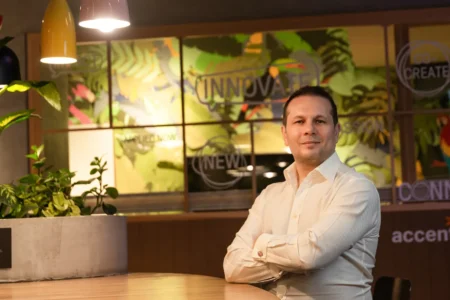 Man in a white shirt sits with arms crossed against a warm, plant-filled office mural, a professional business headshot.