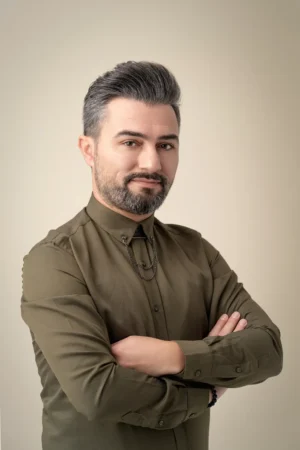 Confident man in an olive shirt with folded arms against a neutral backdrop, studio-lit professional headshot.