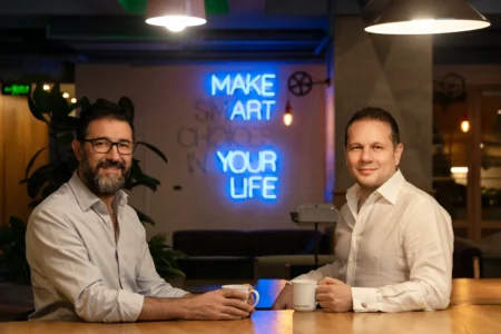 Two smiling men in white shirts sit at a wooden table holding coffee mugs in a warm-lit, authentic team portrait.