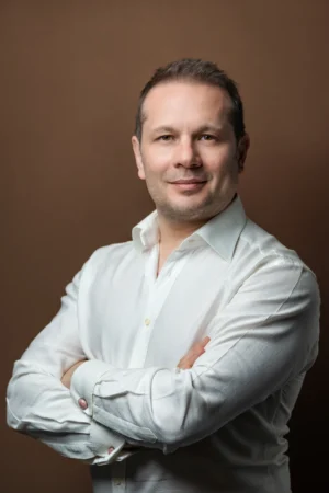 Confident man in a white shirt with arms crossed, studio-lit business headshot against a warm brown backdrop.