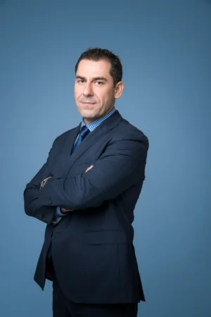 Confident corporate headshot of a man in a navy suit with arms crossed against a smooth blue studio backdrop.
