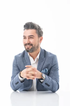 Smiling bearded man in a blue blazer and white shirt, hands clasped on a white background, modern professional headshot.