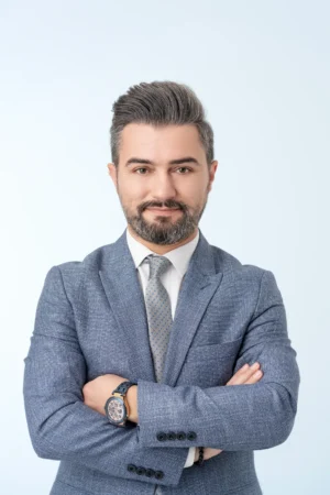 Confident man with salt-and-pepper hair and beard, arms crossed in a blue suit and tie, soft studio-lit professional headshot.