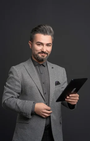 Confident man with salt-and-pepper hair and patterned blazer holding a tablet on a dark studio background, business headshot.