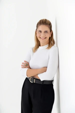 Smiling woman leans against a white wall with arms crossed in a modern professional headshot, soft studio lighting.