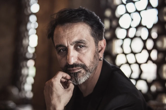 Bearded man in a dark jacket poses thoughtfully against a patterned backdrop with cinematic lighting, professional headshot.