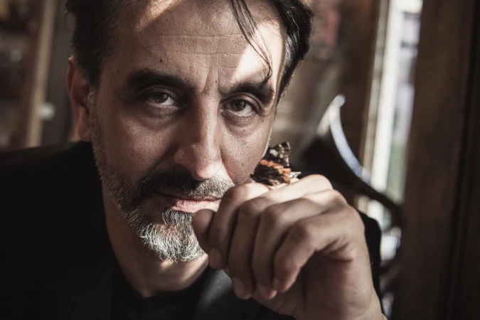 Moody close-up of a bearded man with an intense gaze and a butterfly on his knuckle, cinematic actor headshot.