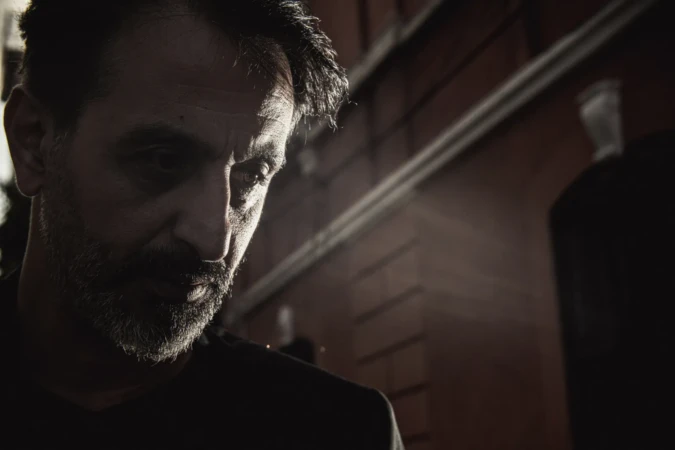 Moody side-lit close-up of a bearded man against a brick wall, a cinematic environmental portrait.
