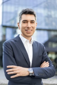 Smiling man in a navy suit poses with arms crossed against a glass office building in a confident business headshot.