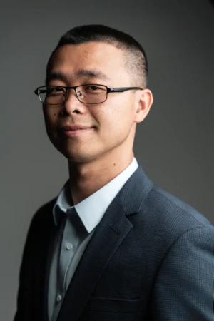 Smiling man in a suit and glasses lit by soft studio light, a confident corporate headshot.