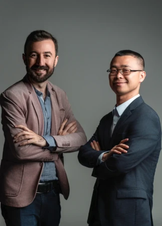 Two smiling men stand with arms crossed against a neutral backdrop in a confident corporate headshot team portrait.