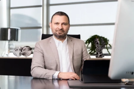 Confident man in a light gray blazer sits at an office desk for a business headshot with soft window light.