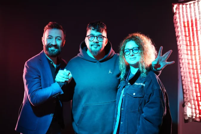 Three musicians pose together in vibrant lighting with a dark backdrop, showcasing camaraderie and creative energy.