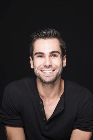 Smiling young man in a black shirt, studio-lit close-up with a dark background, an authentic actor headshot.