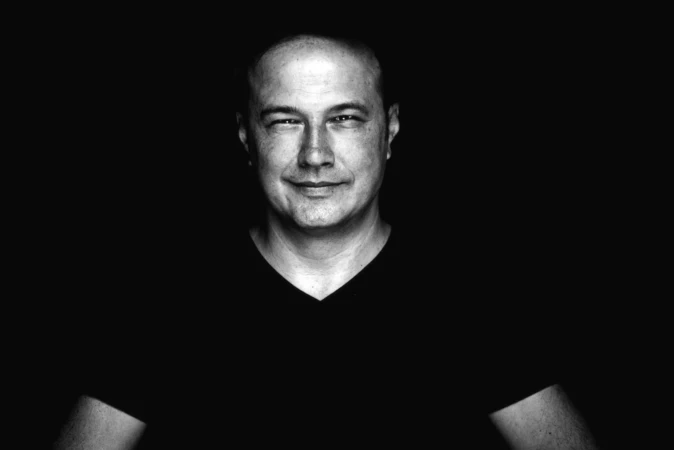 Low-key black-and-white portrait of a man in a V-neck with a confident half-smile, a cinematic professional headshot.
