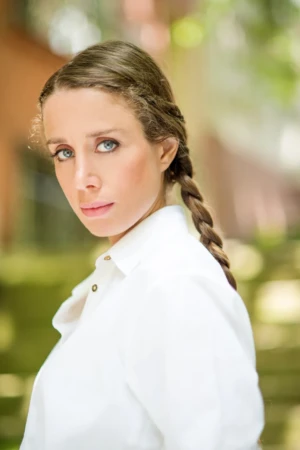 Close-up natural-light professional headshot of a woman with braided hair and white shirt against soft green bokeh.