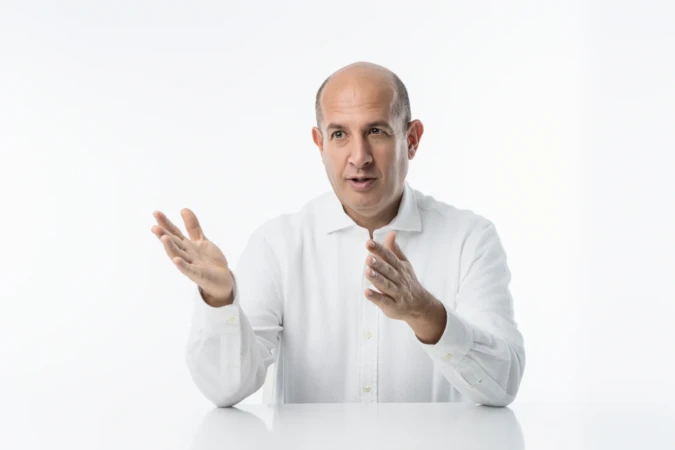 Expressive business headshot of a bald man in a white shirt gesturing confidently against a bright studio background.