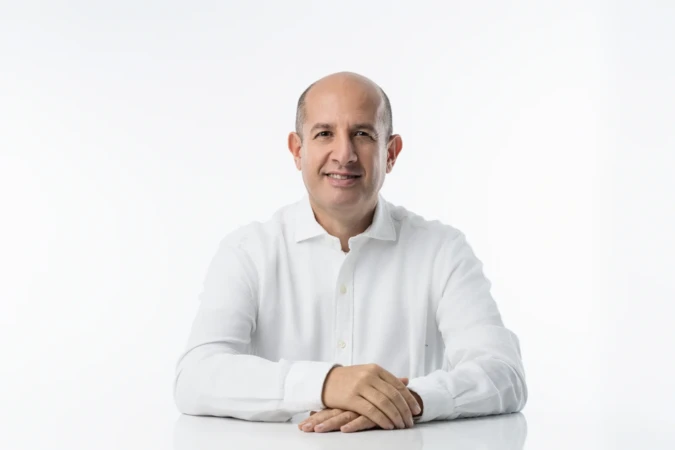 Smiling middle-aged man in a white shirt sits at a white table against a bright backdrop, confident professional headshot.