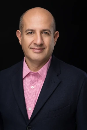 Confident man with a soft studio-lit smile, wearing a pink checked shirt and dark blazer in a professional headshot.