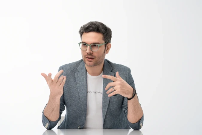 Man in glasses and blazer gestures while speaking against a clean white background in a modern professional headshot.
