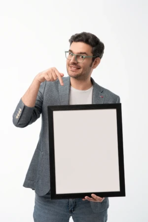 Smiling man in glasses and a grey blazer points at a blank sign against a white backdrop in a modern personal branding photo.