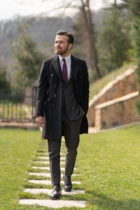 Well-dressed man in a black coat walks confidently on stone pathway with green grass, set against a blurred natural backdrop.