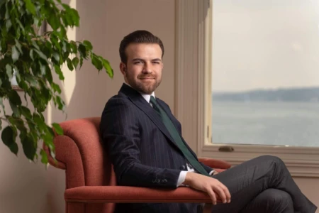 Confident man in a pinstriped suit seated on a bright armchair, framed by greenery and a scenic window view, ideal for business portraits.