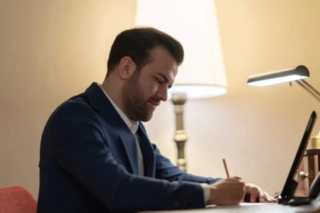 Professional man in a blue suit writing at a desk, illuminated by a desk lamp, with a tablet and warm color backdrop. Perfect for business headshots.