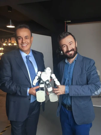 Two smiling men in blue suits hold a white robot in a well-lit studio, an authentic studio portrait.