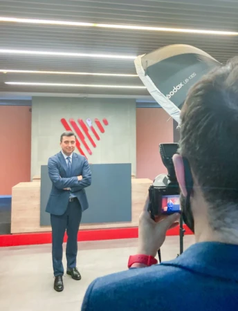 A suited man poses confidently, arms crossed in a reception as a photographer captures a corporate headshot with studio light.