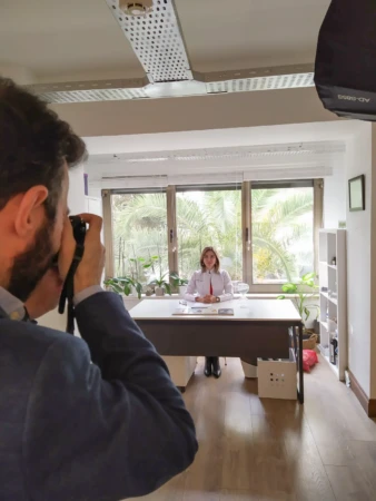 Photographer shooting a seated woman at her desk by a large window with plants, a professional headshot in natural light.