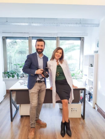 Photographer and smiling doctor in a white coat pose by a sunlit office window, an authentic personal branding photo.