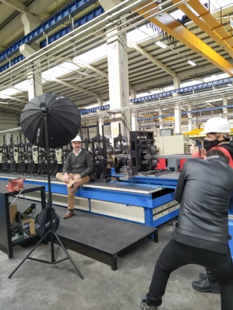 Man in a hard hat sits on factory machinery under softbox light as a photographer captures a modern environmental portrait.