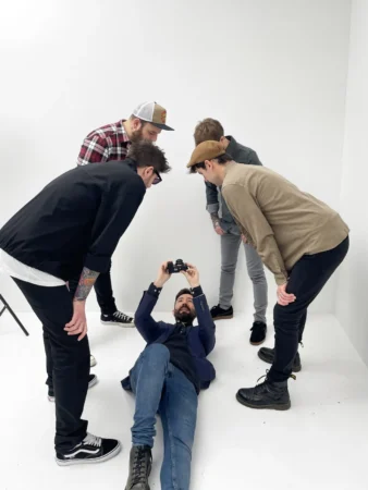 Bearded man lies on white studio floor holding a camera while four casually dressed men lean in, dynamic studio portrait.