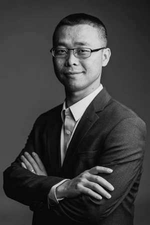 Confident businessman in a suit and glasses poses with arms crossed against a dark backdrop in a professional headshot.